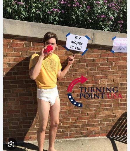Charlie Kirk in a nappy and yellow shirt and drinking from a baby bottle. He's standing in front of a brick wall next to a sign saying "my diaper is full". The lower right corner shows the turning point USA logo .